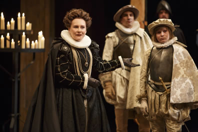 Constance in black dress and cape with silver necklesses, ruff wrists cuffs and collar, and black gloves points to Arthur in armored breast plate, gold patterened cape, ruff collar, pleated baggy pants and gloves and cap, with the Dauphin behind dressed similarly; one of the candalabras is on the left side of the picture.